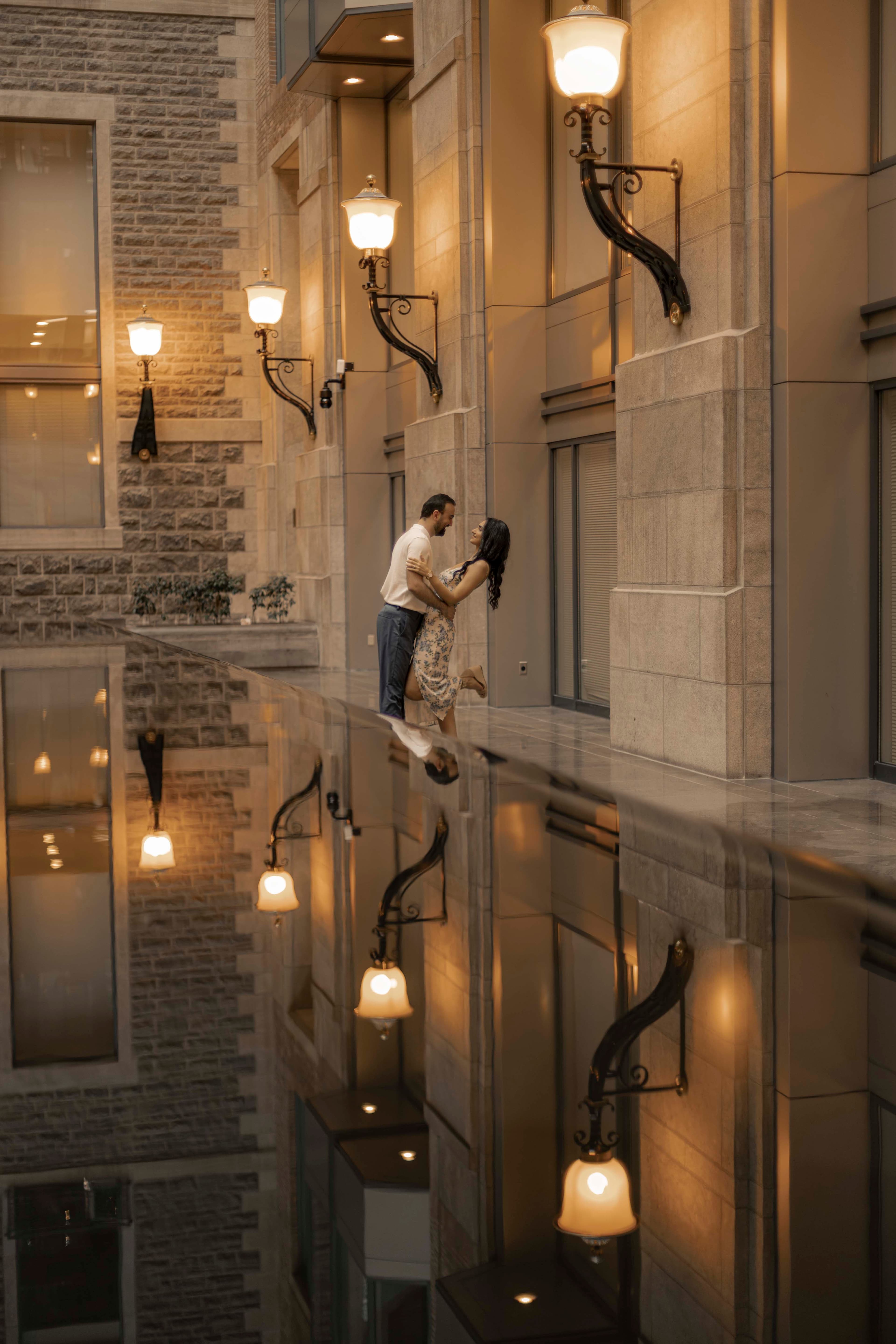 A couple framed in a window, warm sconces along the facade